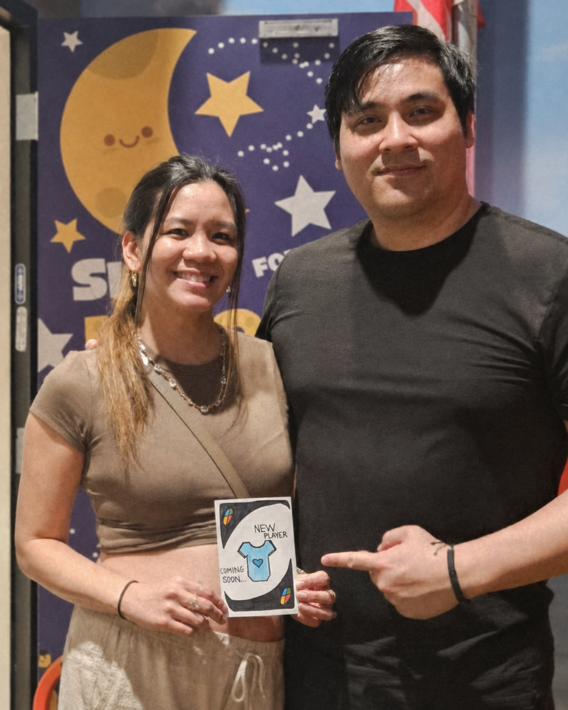 Mikey and Kat holding their New Player Coming Soon card with a blue baby onesie